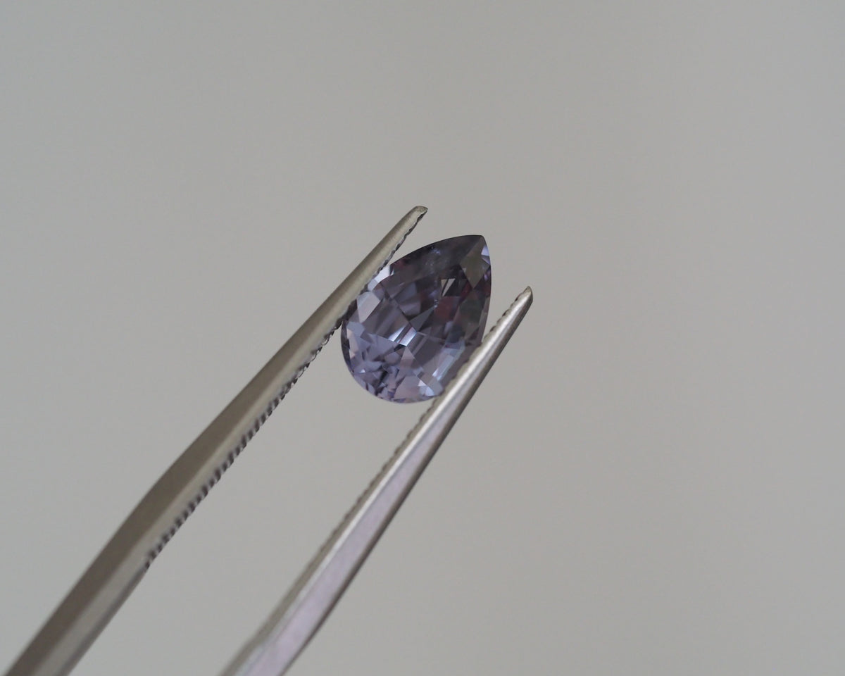 A faceted pear-cut purple gemstone held between silver metal tweezers against a plain light gray background.