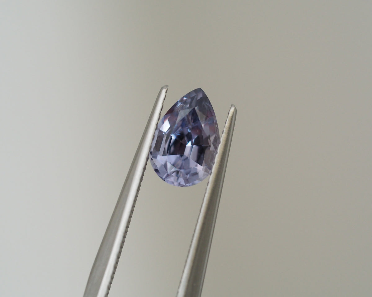 A teardrop-shaped faceted lavender gemstone held in the jaws of silver metal tweezers against a plain light gray background.
