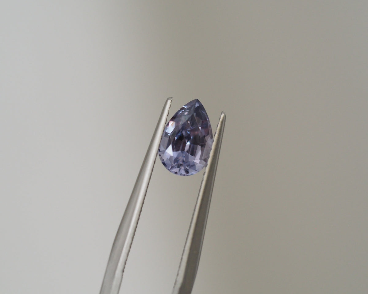 A teardrop-shaped light purple gemstone held in the jaws of metal tweezers against a plain light gray background.