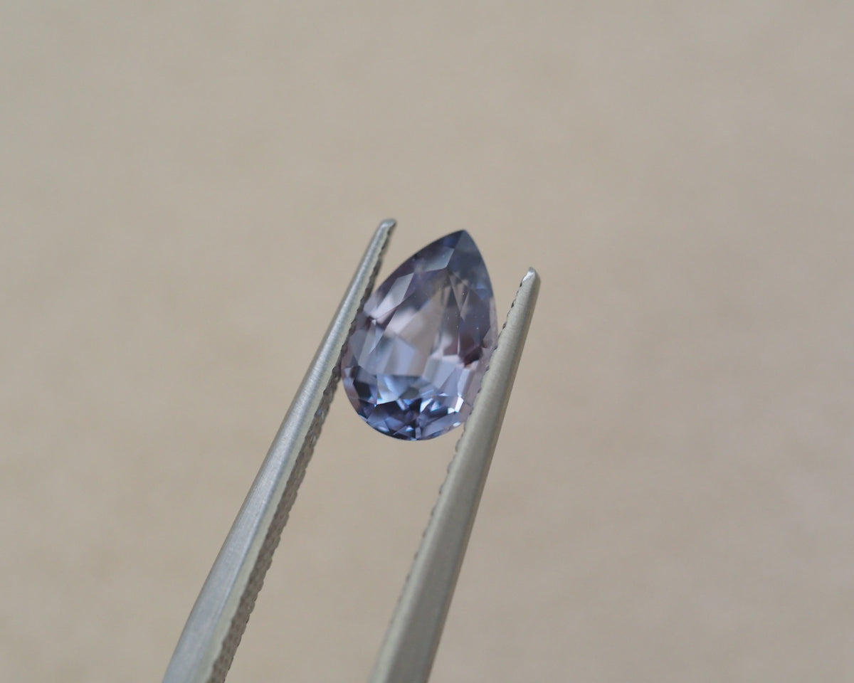 A faceted pear-shaped gemstone in a pale lavender-blue hue, held between metal tweezers against a plain beige background.