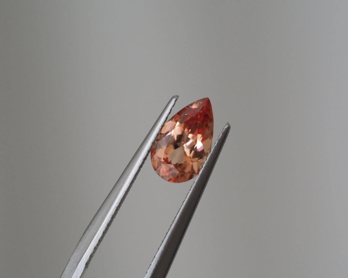 A faceted pear-shaped gemstone in a peachy orange-red hue, held between silver metal tweezers against a smooth light gray background.