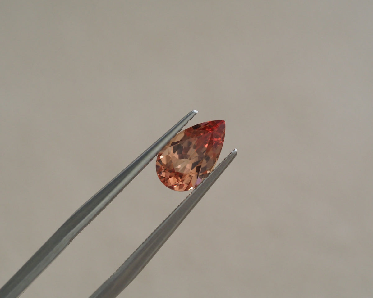 A teardrop-shaped peach-colored gemstone held between silver tweezers against a plain beige background.