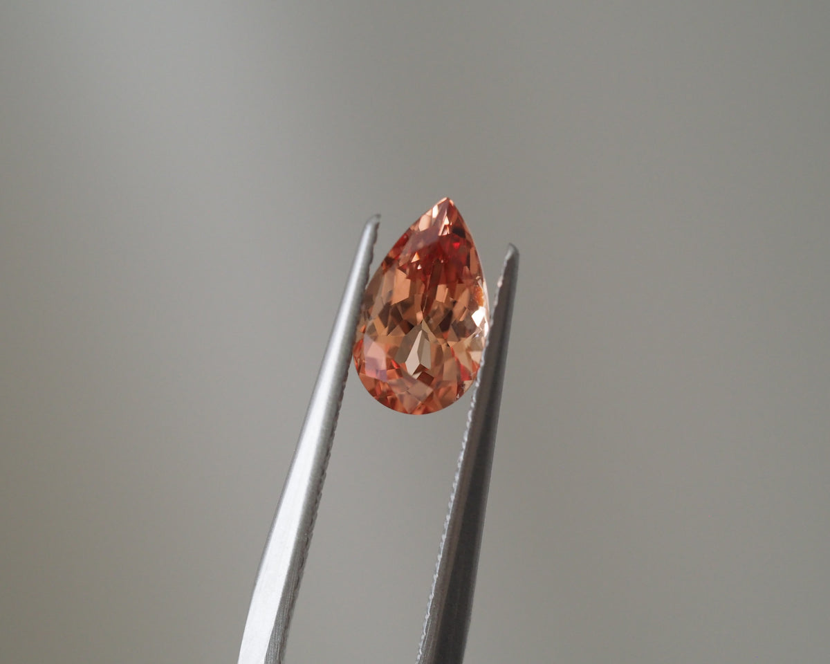 A teardrop-shaped peach-colored gemstone held between metal tweezers against a soft gray background.