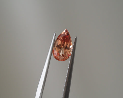 A teardrop-shaped peach-colored gemstone held between metal tweezers against a soft gray background.