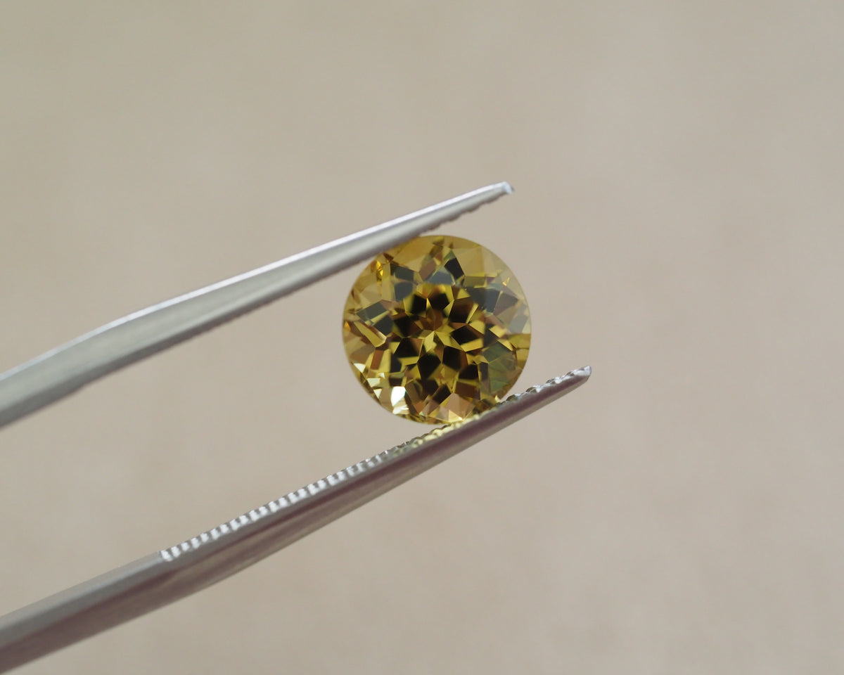 A round yellow gemstone held between silver metal tweezers against a soft beige background.