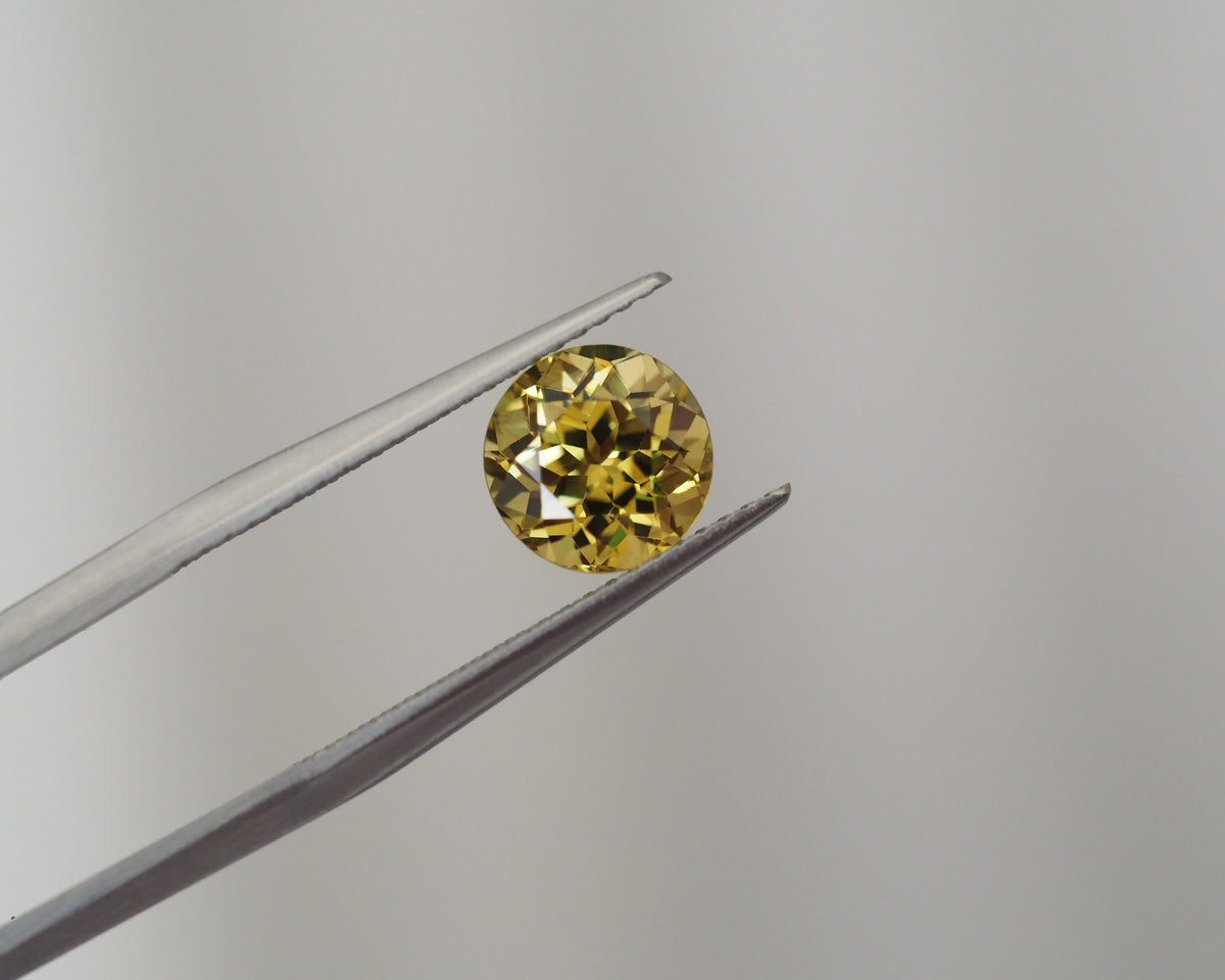 A round yellow gemstone held between metal tweezers against a plain light gray background.