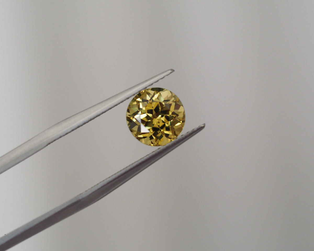 A round yellow gemstone held between metal tweezers against a plain light gray background.