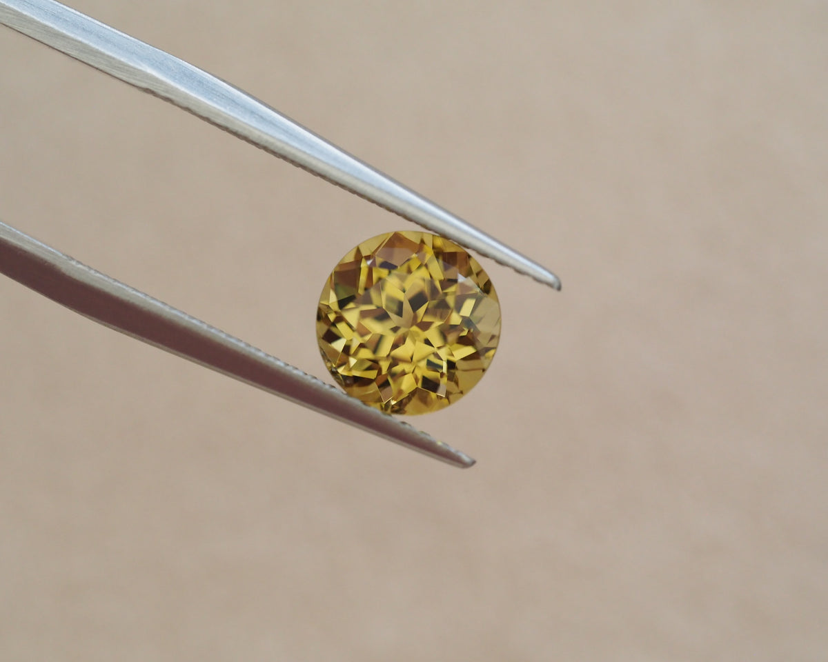 A round yellow gemstone held between metal tweezers against a plain beige background.