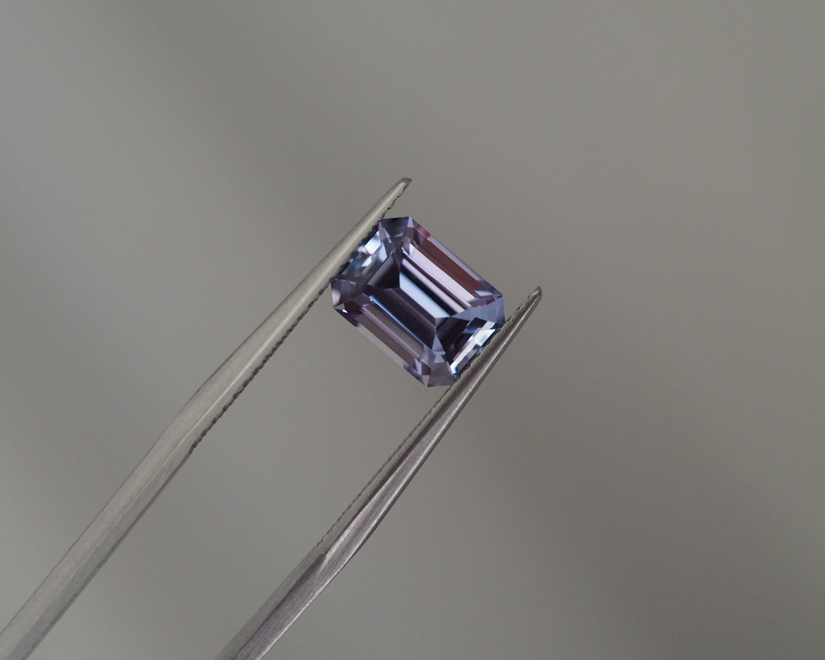 A rectangular emerald-cut lavender gemstone held between metal tweezers against a soft gray background.