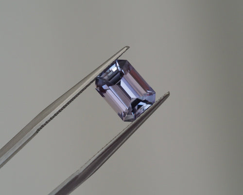 A loose rectangular emerald-cut lavender gemstone held between metal tweezers against a plain light gray background.
