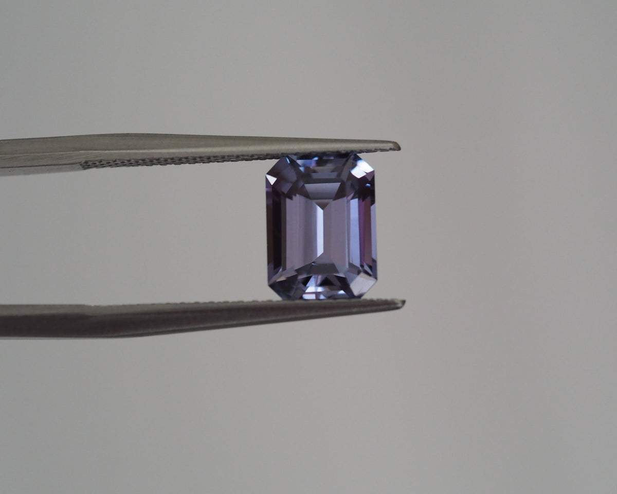 A loose rectangular emerald-cut purple gemstone held between metal tweezers against a plain light gray background.