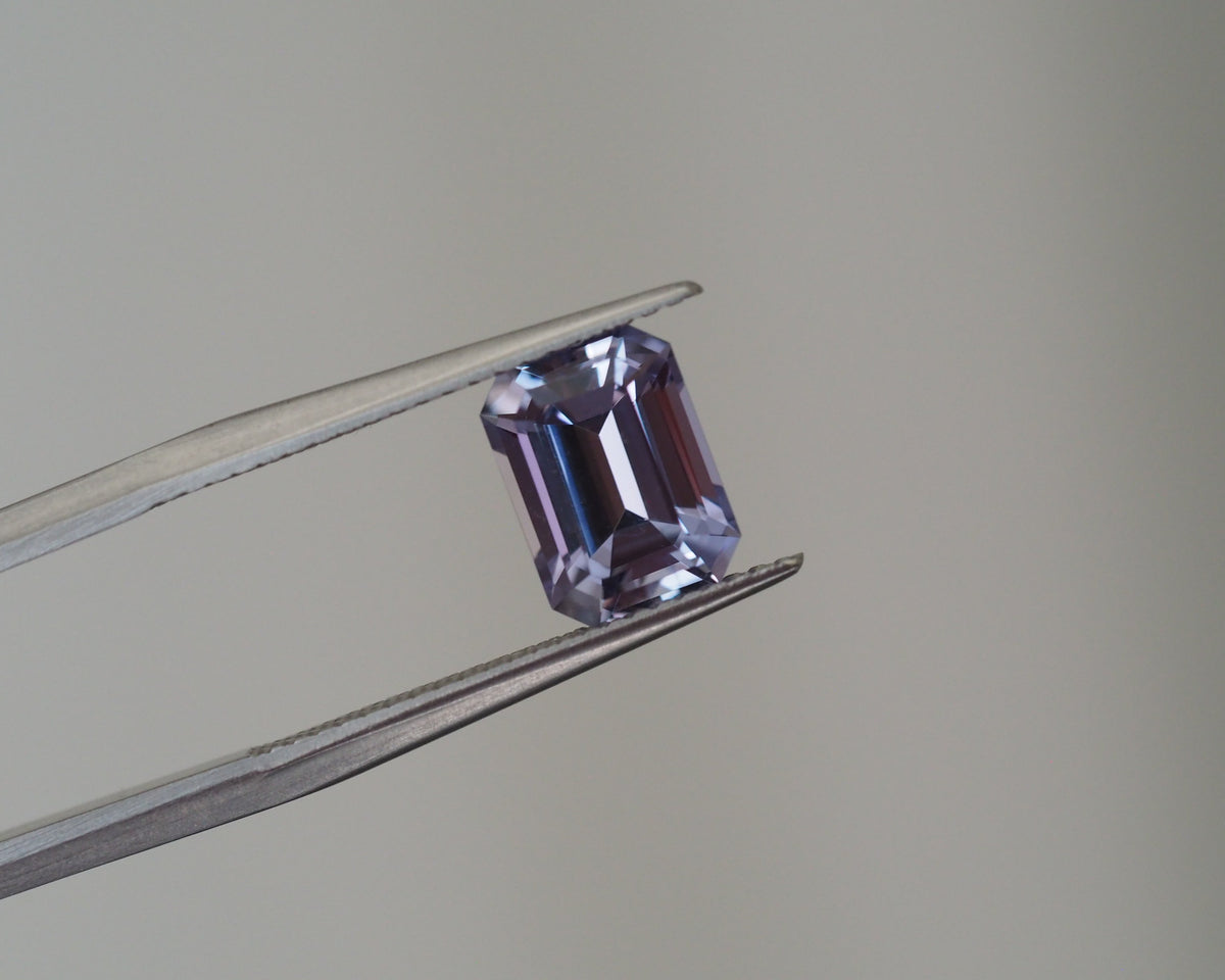 A rectangular emerald-cut purple gemstone held in silver tweezers against a neutral gray background.