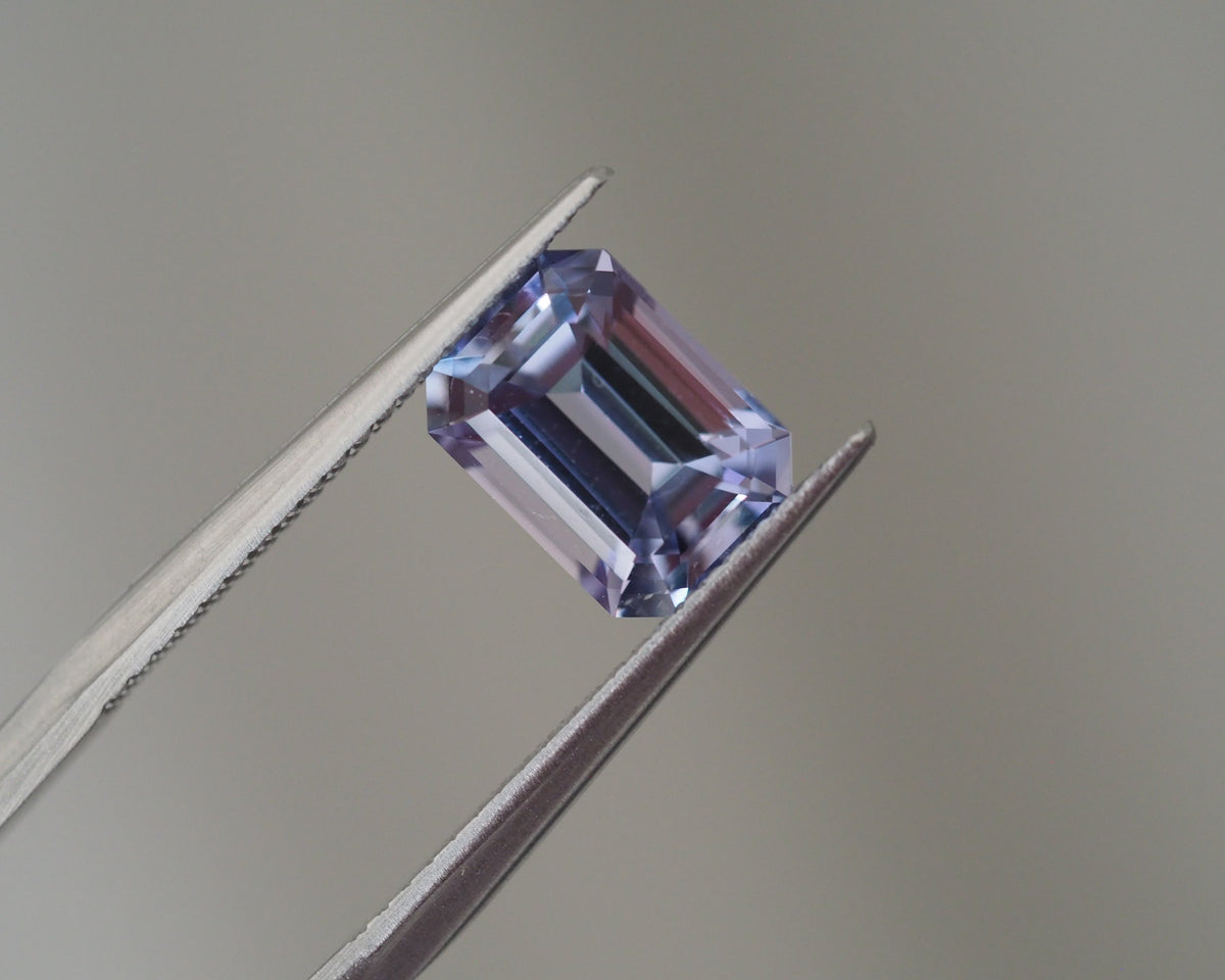 A rectangular emerald-cut purple gemstone held between metal tweezers against a soft gray background.