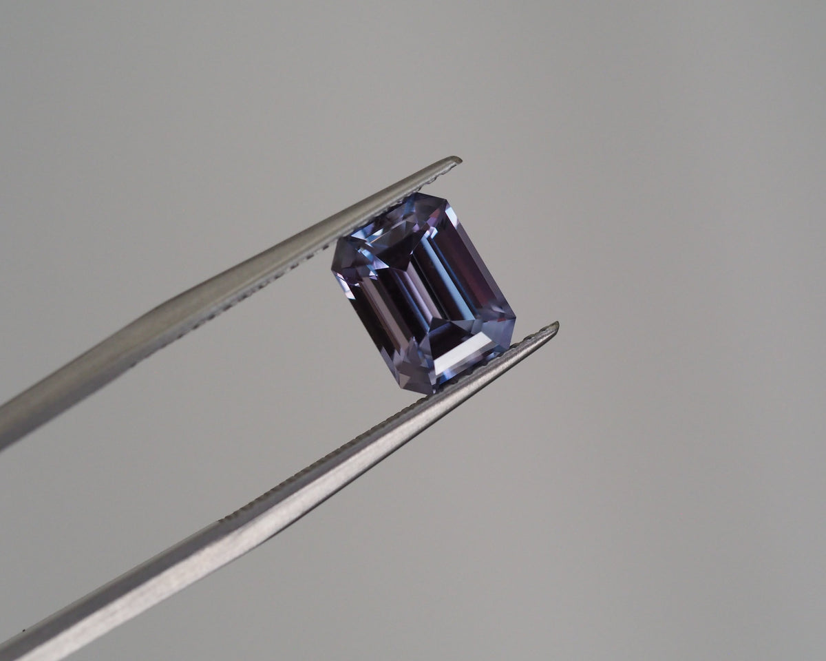 A loose emerald-cut purple gemstone held between metal tweezers against a plain light gray background.