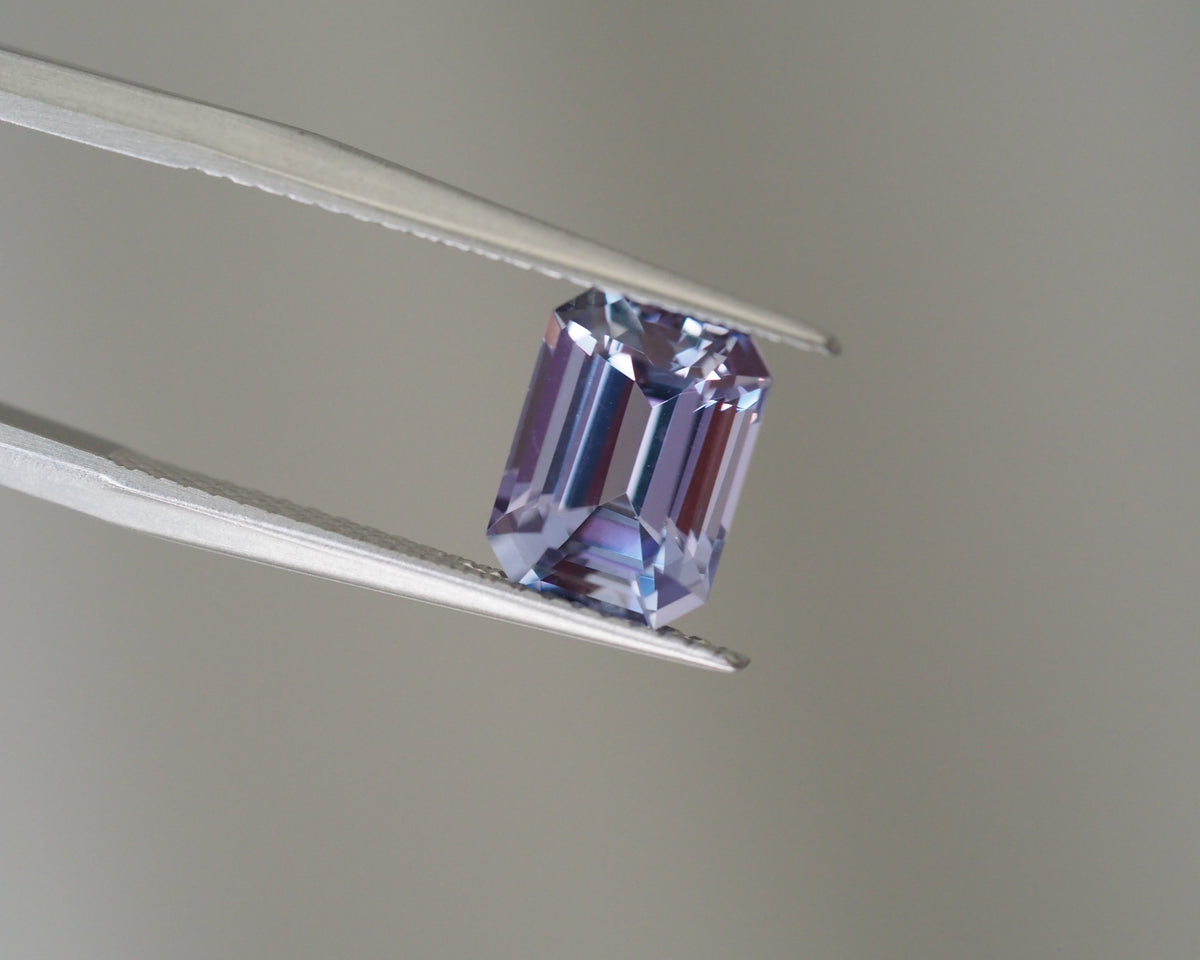 A loose emerald-cut lavender purple gemstone held between silver metal tweezers against a neutral gray background.