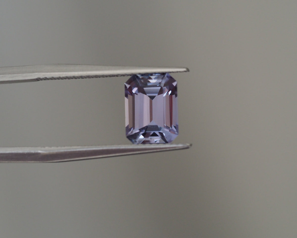 An emerald-cut lavender purple tanzanite held between silver metal tweezers against a soft gray background.