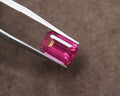 A rectangular emerald-cut fuschia pink rhodolite held between silver metal tweezers against a textured brown background.