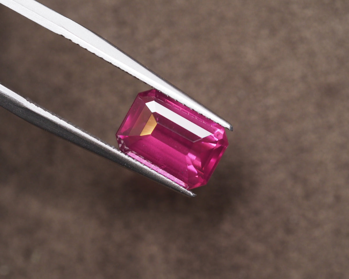 A rectangular emerald-cut fuschia pink rhodolite held between silver metal tweezers against a textured brown background.