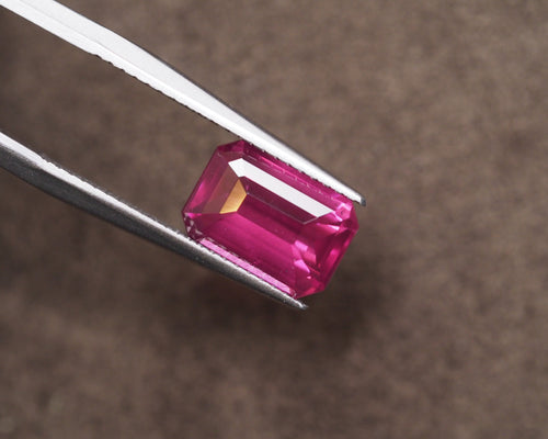 A rectangular emerald-cut fuschia pink rhodolite held between silver metal tweezers against a textured brown background.