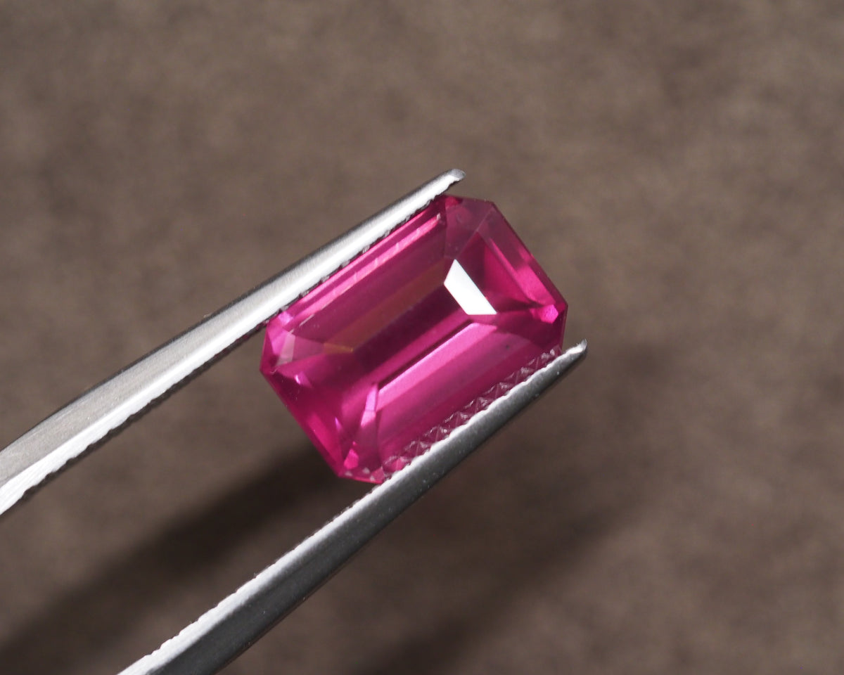 A bright pink rectangular emerald-cut Rhodolite held between silver metal tweezers against a soft brown textured background.