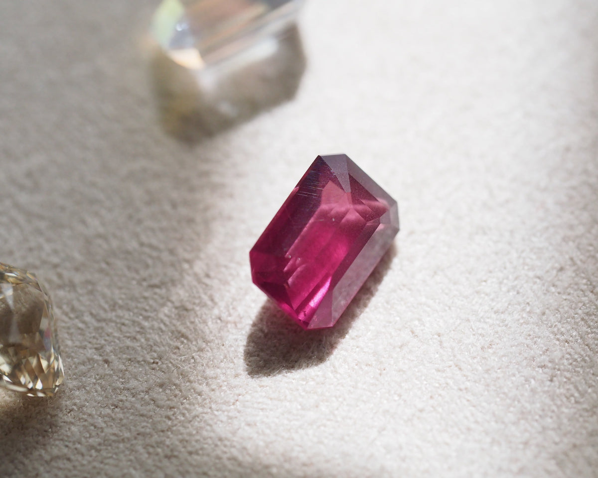 A faceted rectangular pink rhodolite with a brilliant cut, displayed on a textured beige surface next to two clear, uncut gemstones under warm directional light.