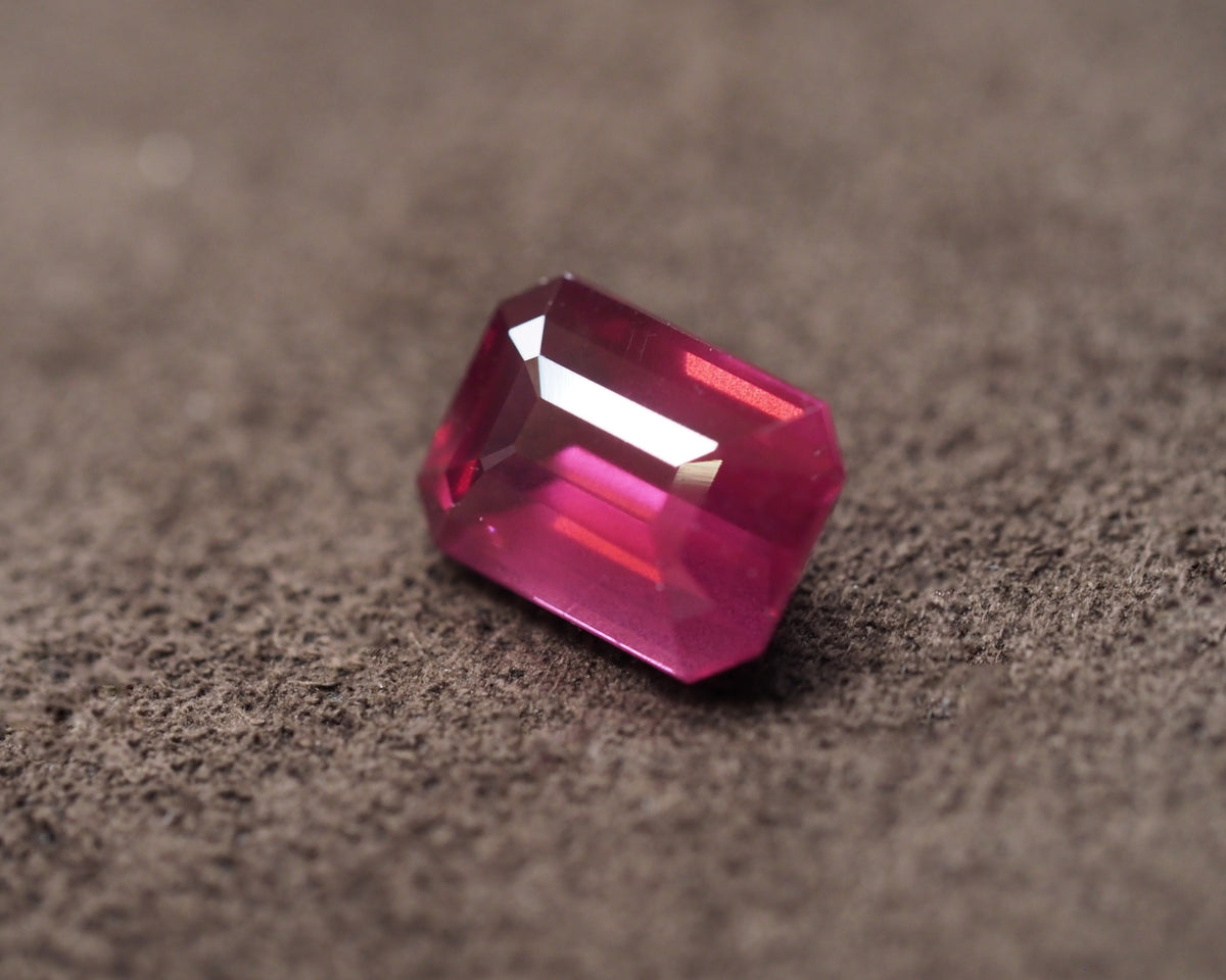 A faceted rectangular hot pink rhodolite with a emerald cut, resting on a textured dark gray surface.
