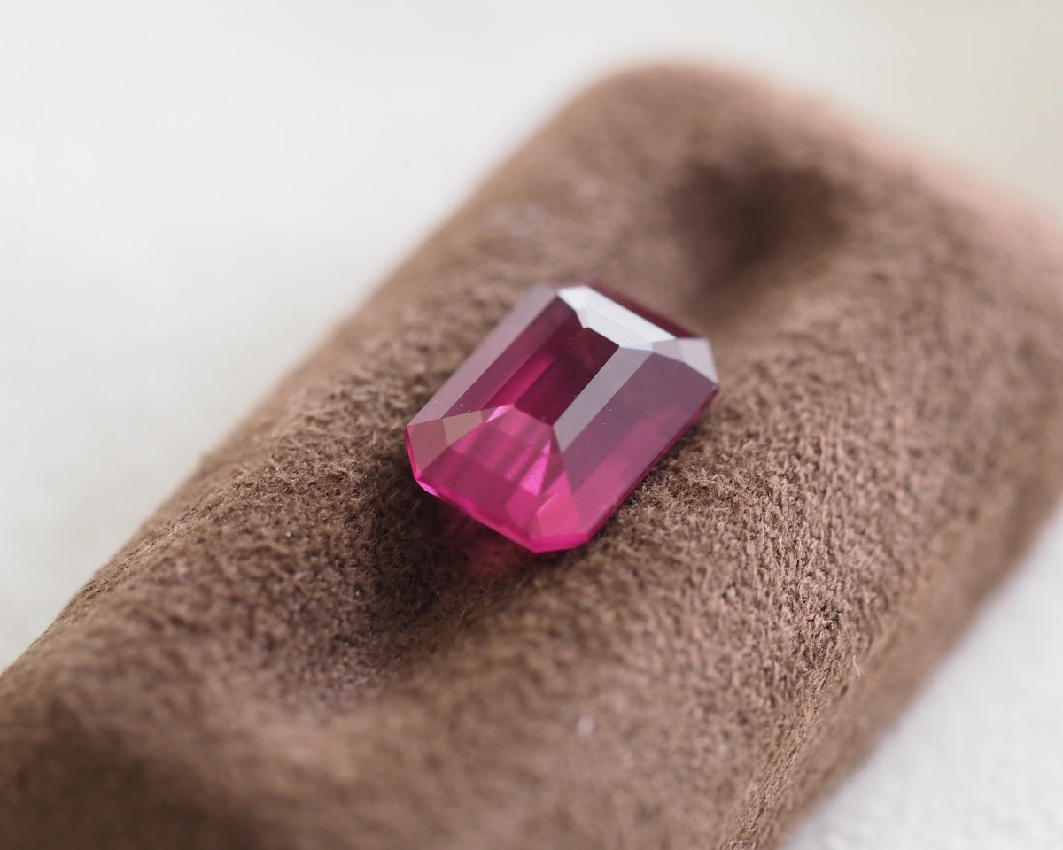 A faceted rectangular pink gemstone, likely a ruby or garnet, displayed on a textured brown velvet jewelry stand against a soft, out-of-focus light background.