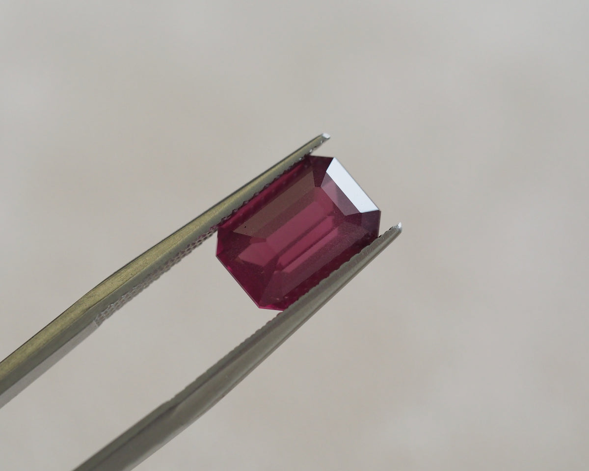 A rectangular emerald-cut deep pinkish-red gemstone held in silver metal tweezers against a soft, light beige background.