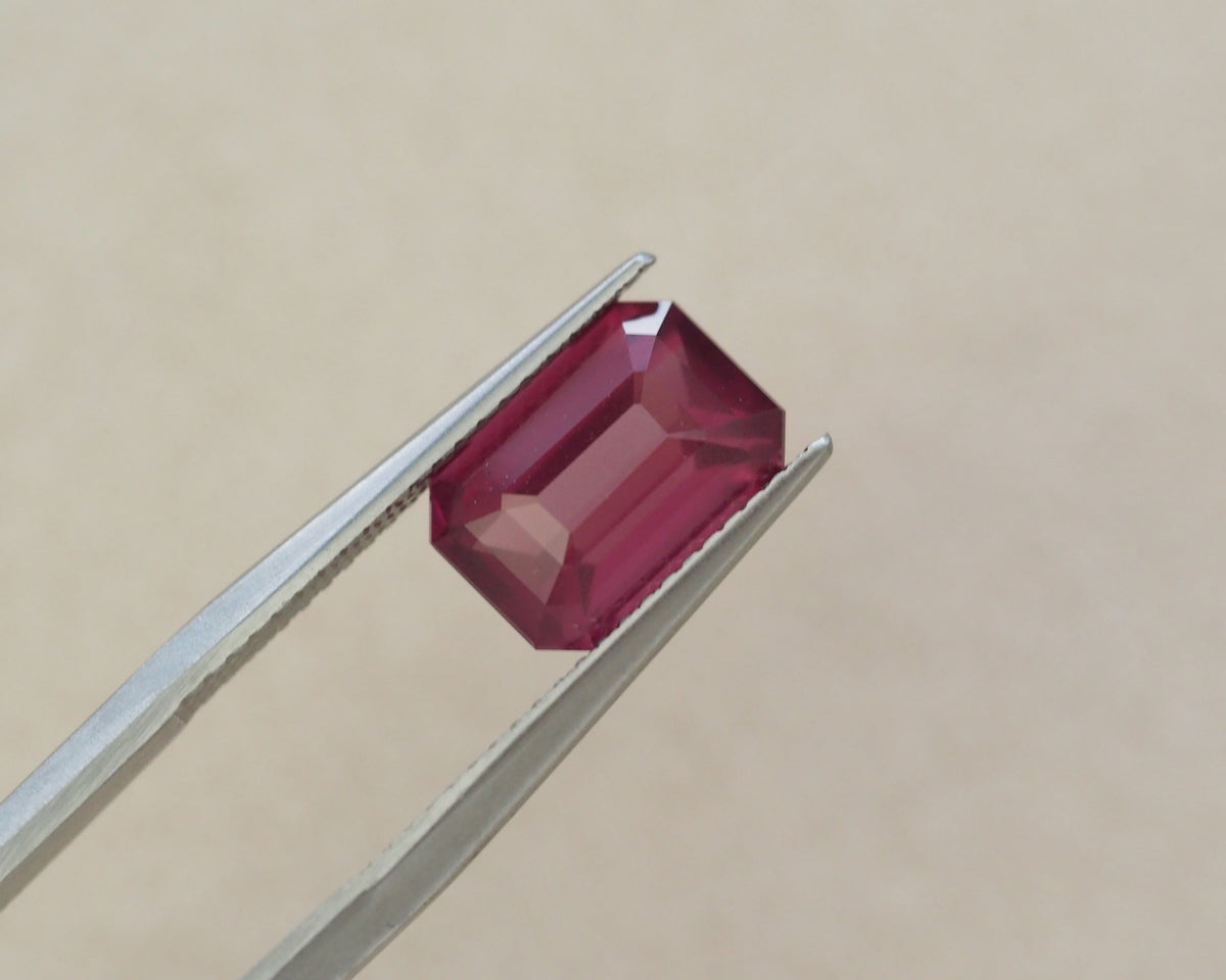 A rectangular emerald-cut pink gemstone held between silver metal tweezers against a plain beige background.