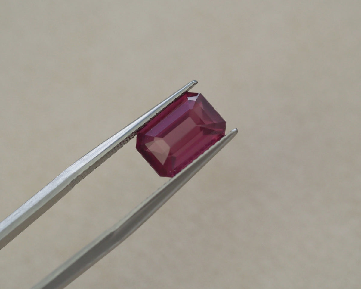 A rectangular emerald-cut pink gemstone held between silver metal tweezers against a plain beige background.