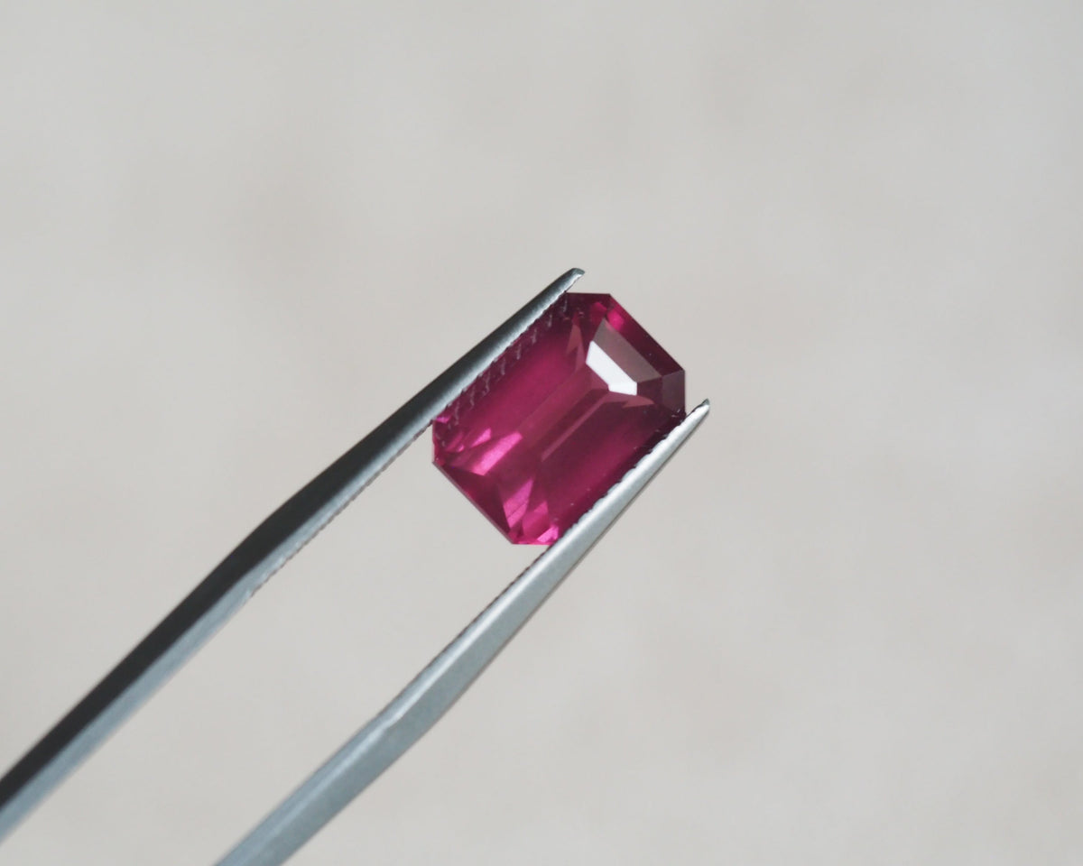 A rectangular emerald-cut pink rhodolite held between metal tweezers against a soft light gray background.