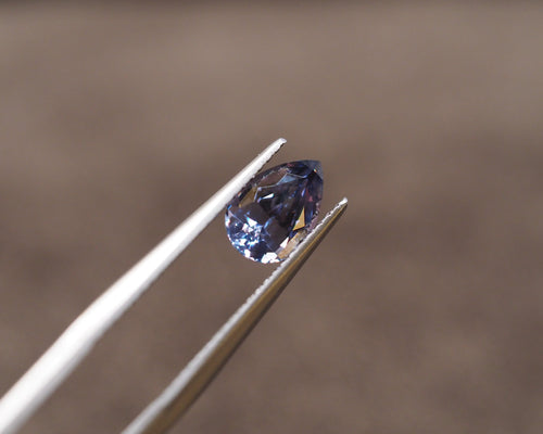 A faceted oval-cut blue gemstone held between metal tweezers against a soft, blurred brown background.