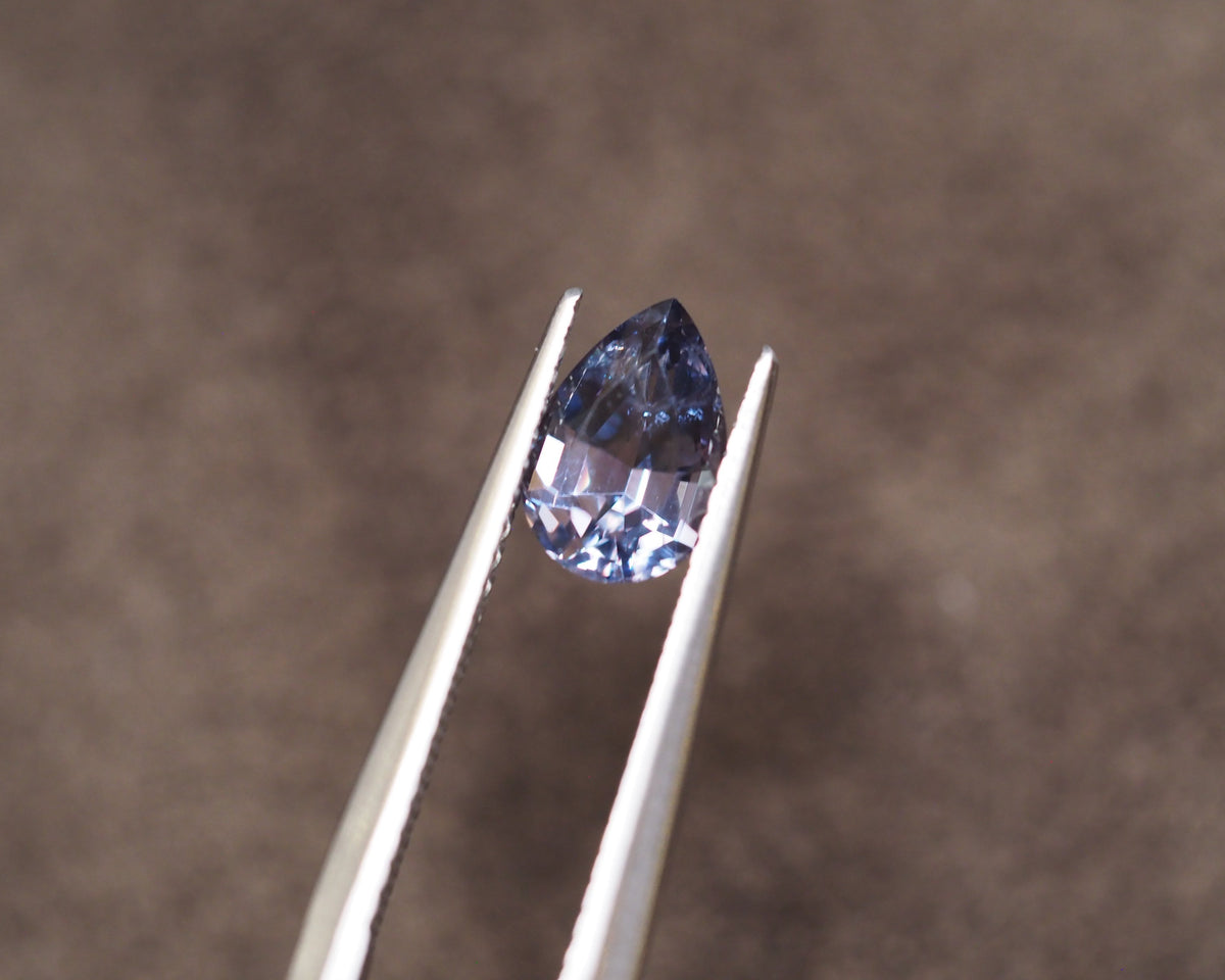 A faceted pear-cut blue gemstone held between silver metal tweezers against a soft, out-of-focus brown background.