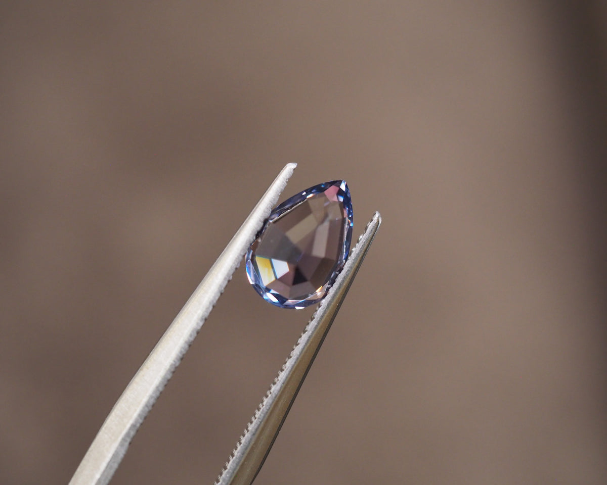 A faceted teardrop-shaped gemstone held between metal tweezers against a soft, blurred brown background.