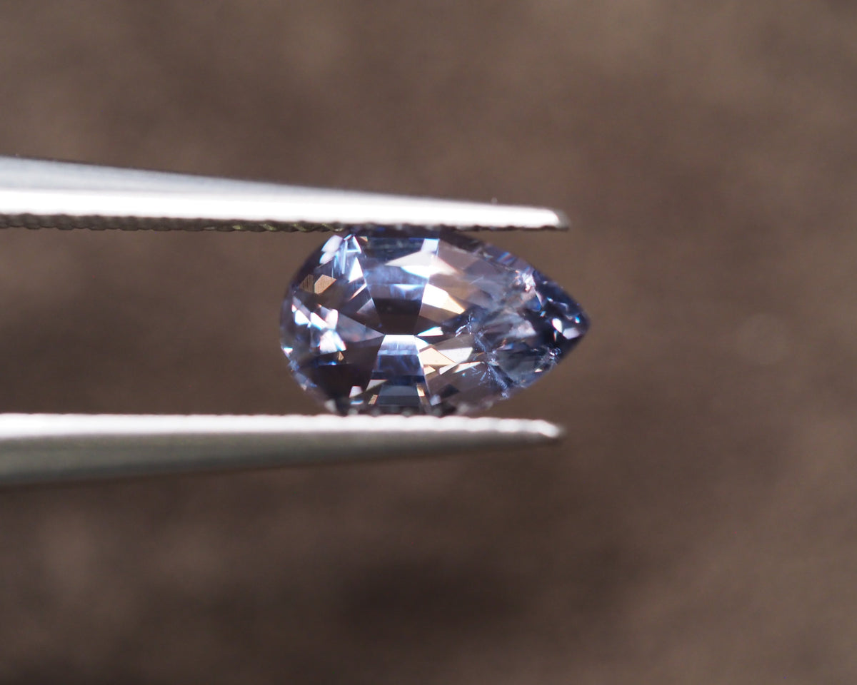 A marquise-cut light blue gemstone held between metal tweezers against a soft, blurred brown background.