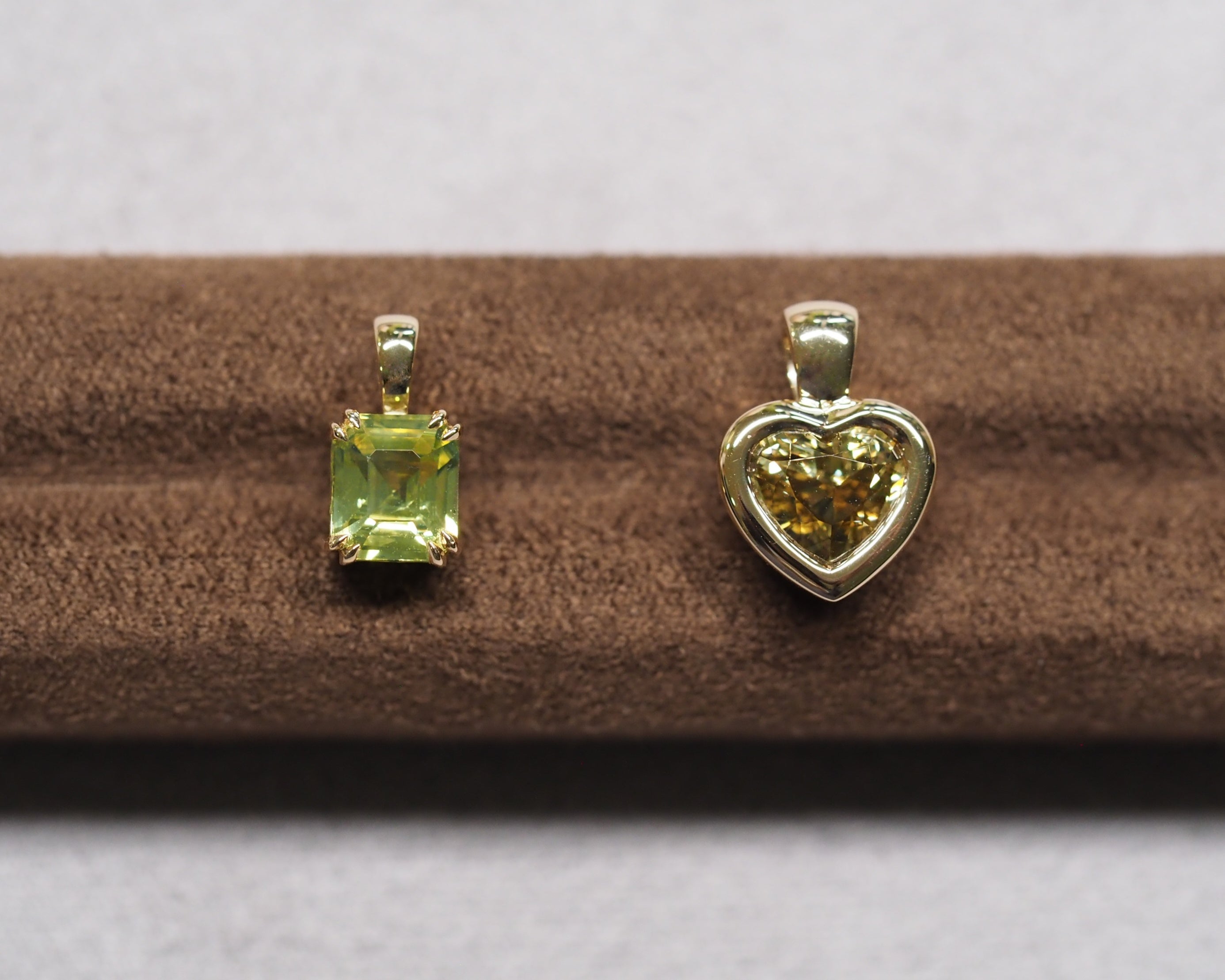 Two yellow gold pendants displayed on a brown velvet jewelry stand against a light gray background. The left pendant features a rectangular green gemstone, and the right pendant features a heart-shaped yellow gemstone.