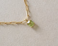 A close-up of a gold paperclip chain necklace with a small square-cut light green gemstone pendant, resting on a textured beige surface.