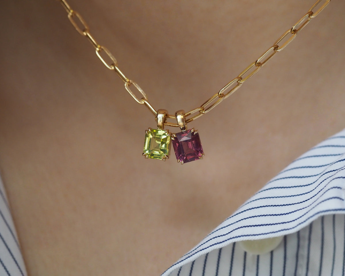 A close-up of a gold paperclip chain necklace worn on a person's neck, featuring two small square gemstone pendants: one light green and one dark pinkish-red, with a blue and white striped shirt visible at the bottom right.