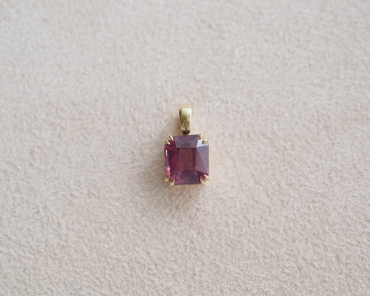 A rectangular pink spinel pendant set in a gold bezel, displayed on a textured light beige surface.