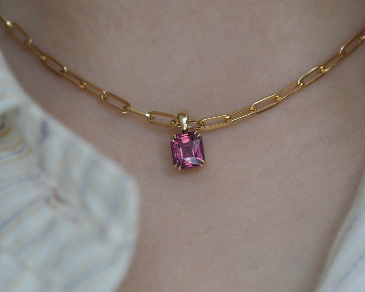 A close-up of a gold paperclip chain necklace with a rectangular pink spinel pendant, worn on a person's neck.