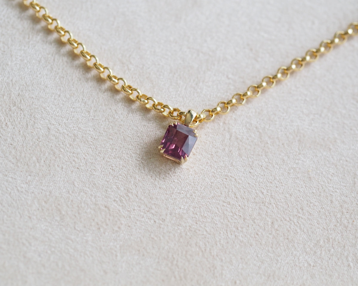 A delicate gold chain necklace featuring a small, square-cut pinkish purple spinel pendant in a gold bezel setting, displayed on a textured cream-colored surface.