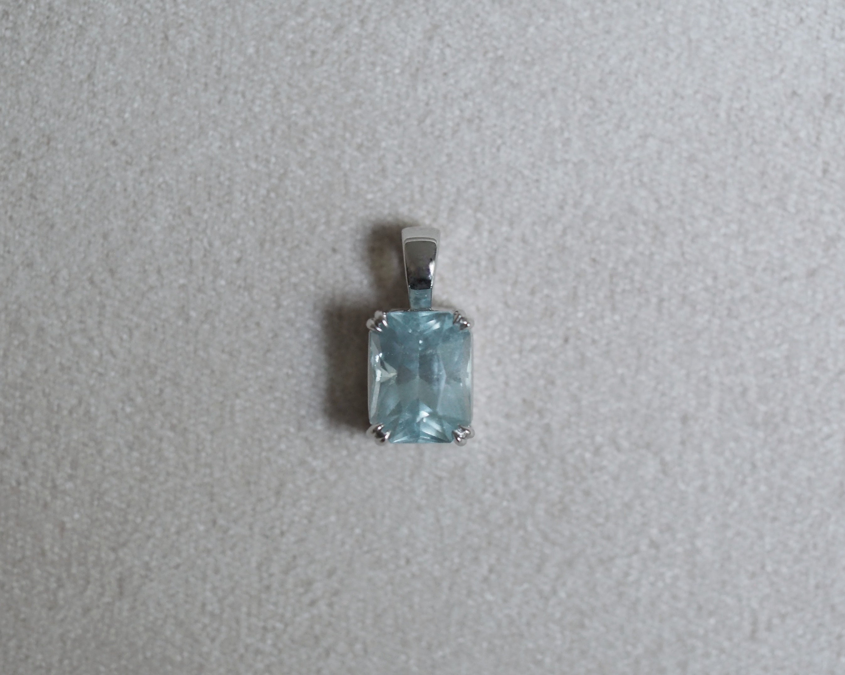 A rectangular light blue gemstone pendant set in a silver-toned metal bezel, shown on a textured light gray background.