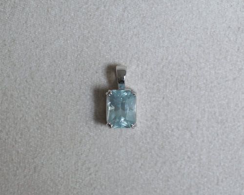A rectangular light blue gemstone pendant set in a silver-toned metal bezel, shown on a textured light gray background.