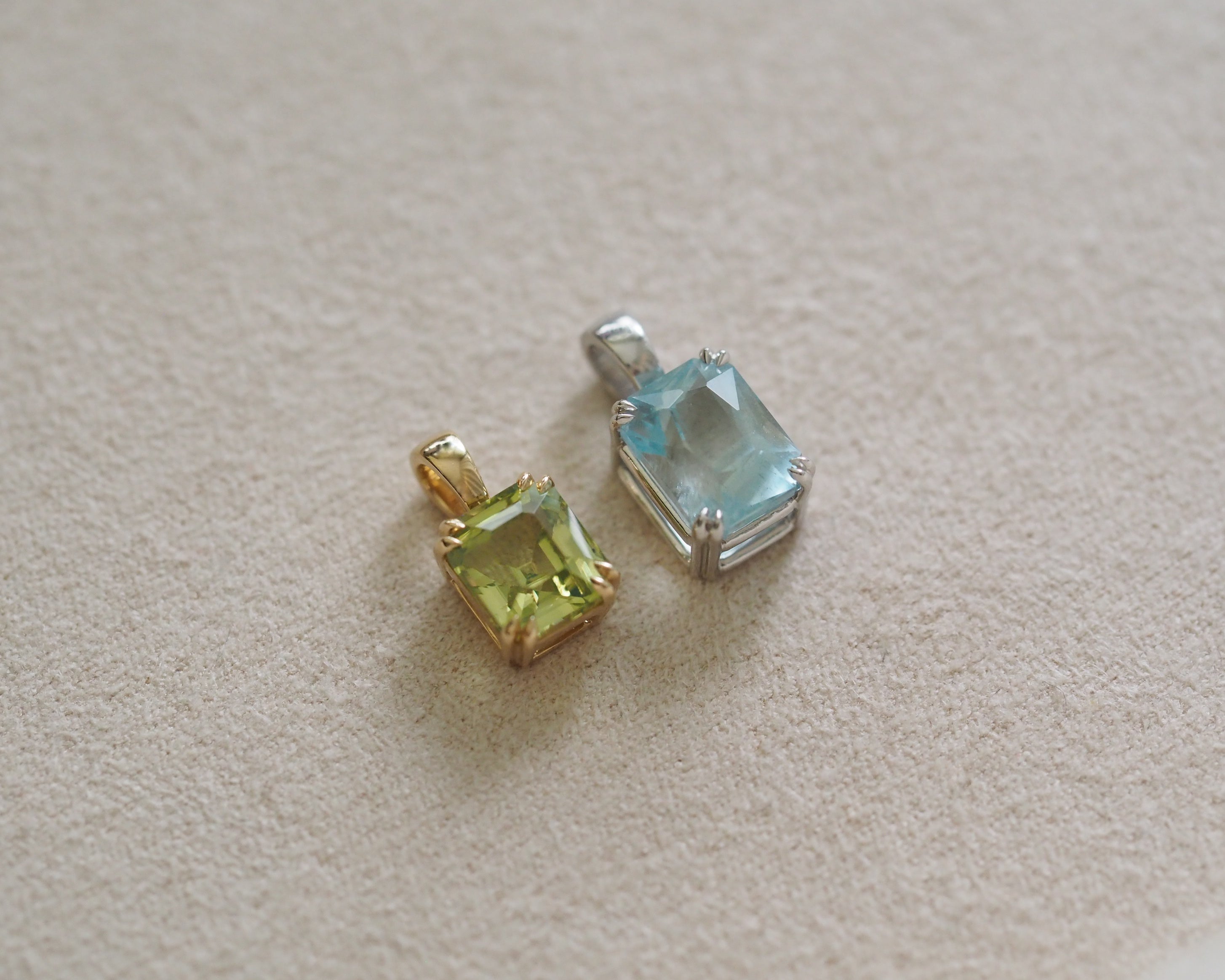 Two octagonal shape gemstone pendants, one in yellow gold and one in silver, displayed on a textured beige surface.