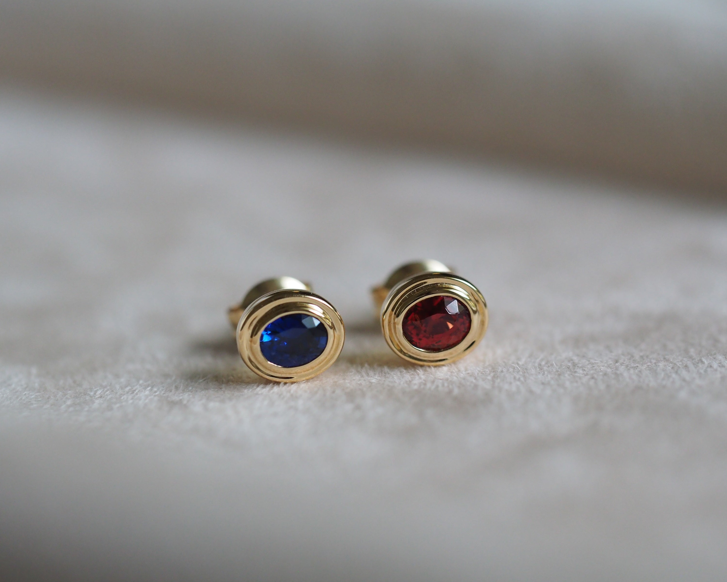 A pair of gold stud earrings with oval-cut blue and red gemstones, resting on a soft, light beige textured surface.