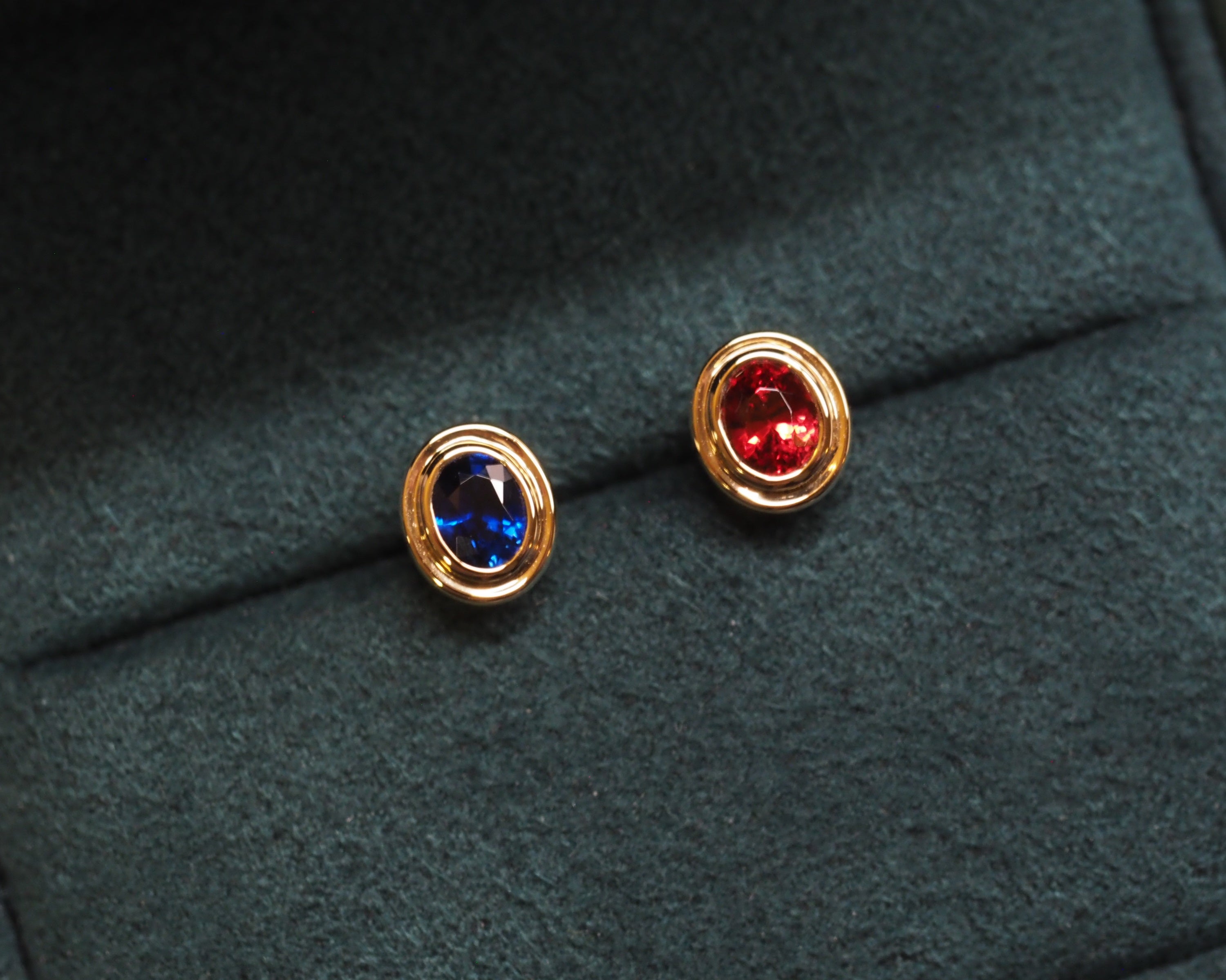 A pair of gold stud earrings with round blue and red gemstones, displayed on a dark green velvet jewelry box.