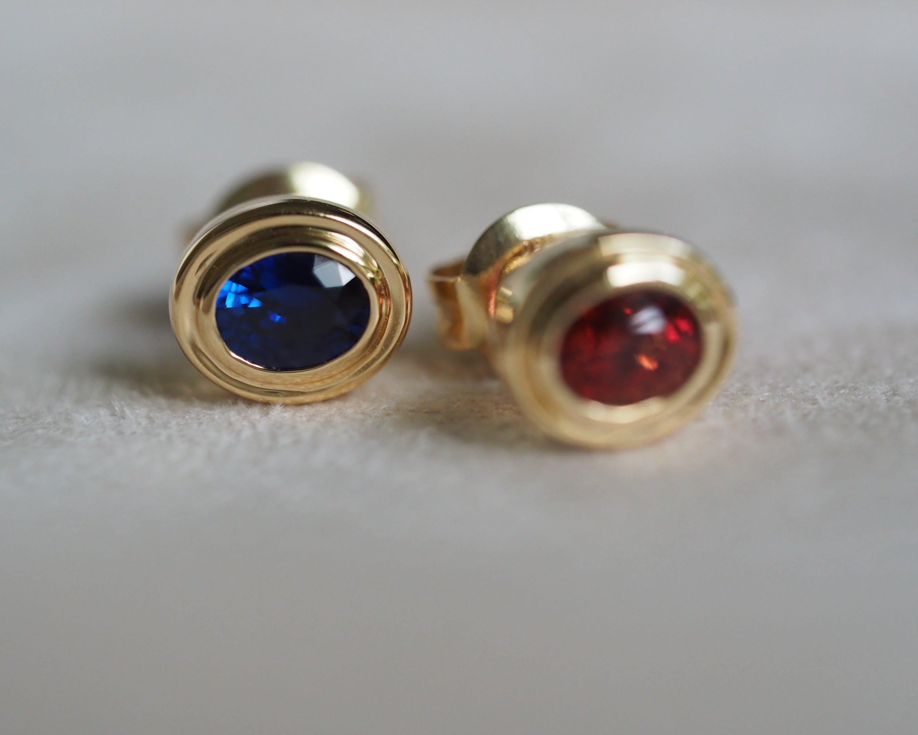 A pair of gold stud earrings with oval-cut blue sapphire and red spinel, displayed on a soft, light gray textured surface.