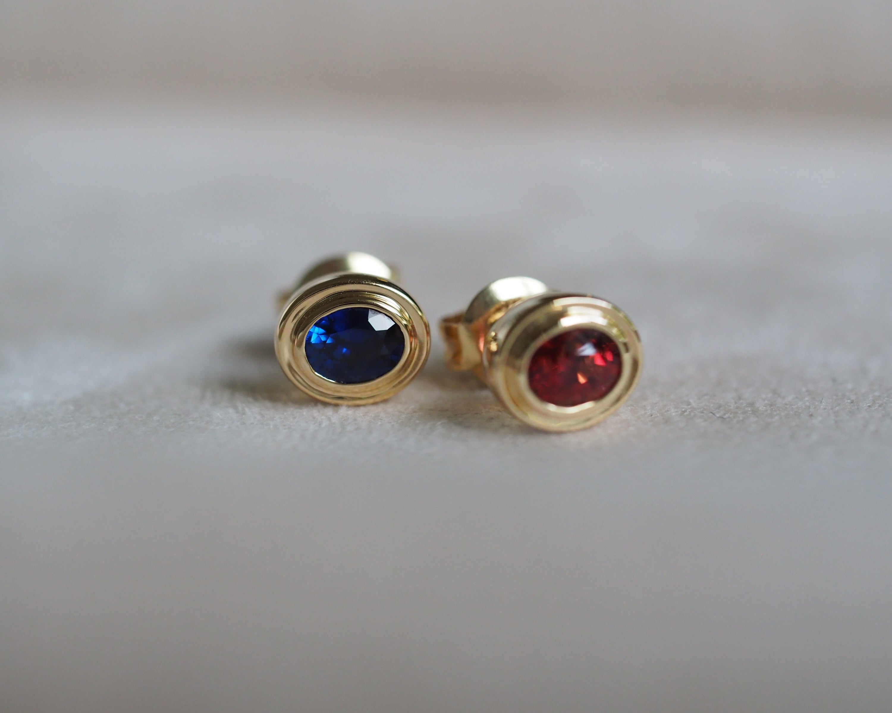 A pair of gold stud earrings with oval-cut blue sapphire and red vanadium spinel, displayed on a soft, light gray fabric surface.