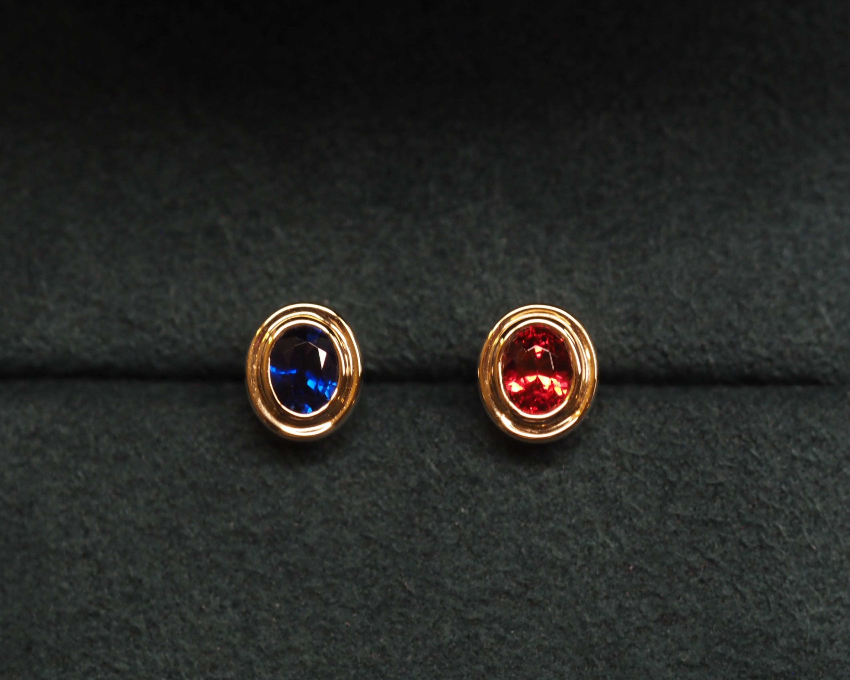 A pair of gold oval stud earrings, one featuring a deep blue gemstone and the other a red gemstone, displayed on a textured black background.