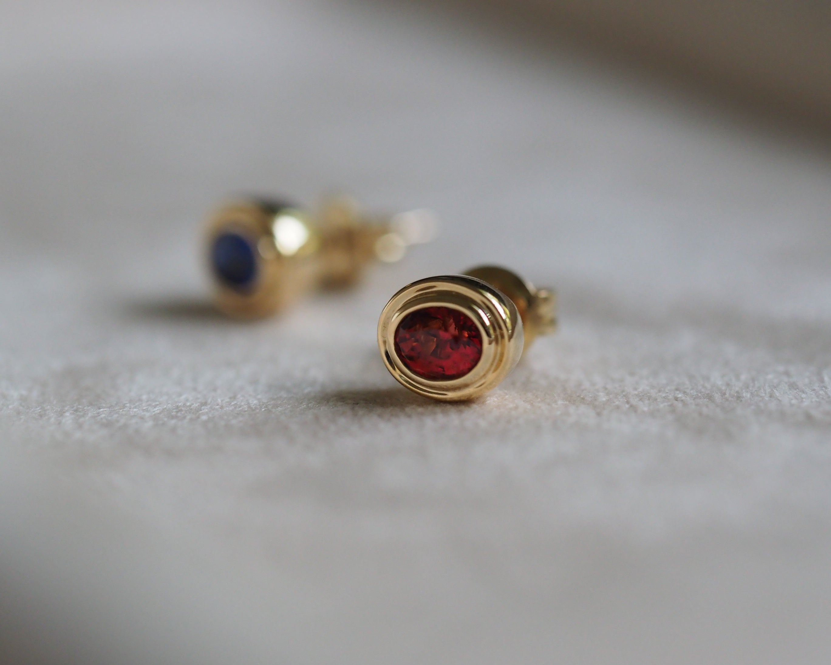 A pair of gold stud earrings with oval gemstones, one in red spinel and one in royal blue sapphire, resting on a soft, light-colored textured surface.
